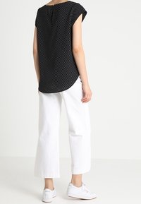 Sort bluse med polkaprikker og korte ærmer, buet søm, parret med hvide brede bukser og hvide sneakers. Enkel design og struktur.