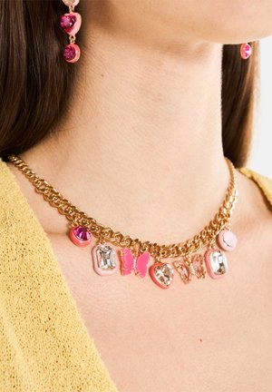 Goldene Kettenhalskette mit pinken und klaren dekorativen Charms, darunter Schmetterlinge und verschiedene Formen, vor einem hellen, strukturierten Stoff.