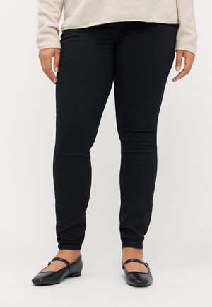 Kvinde iført sorte skinny bukser, beige sweater og sorte Mary Jane-sko, stående mod en ensfarvet hvid baggrund.