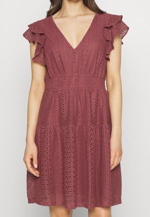 Frau trägt ein knielanges Spitzenkleid in staubigem Rosaton mit Flügelärmeln, V-Ausschnitt, Knopfdetail und elastischem Taillenbund.