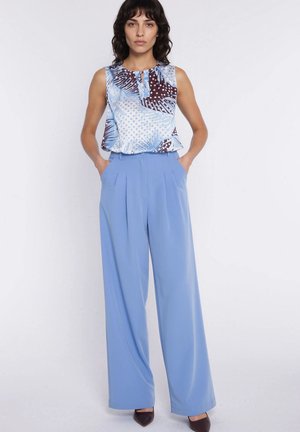 Kobieta stojąca z rękami w kieszeniach, ubrana w bezrękawnik w niebiesko-brązowy wzór oraz szerokie jasnoniebieskie spodnie i ciemne obcasy.