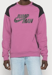 Uomo che indossa una felpa viola e nera con la scritta "JUMPMAN" e un logo a swoosh, abbinata a jeans azzurri.