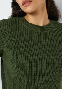 Olivenfarget ribbestrikket genser med rund hals. Teksturen er fremtredende, med vertikale linjer som går gjennom stoffet.