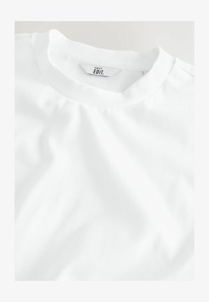 Egyszerű fehér póló, közelről bemutatva a nyakkivágást és a címkét, amelyen a "NEXT EDIT." felirat látható egy világos háttéren.