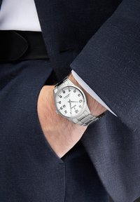 Montre en métal argenté avec un cadran blanc rond, chiffres noirs, affichage de la date et un bracelet en acier inoxydable, portée sur un poignet dans un costume sombre.