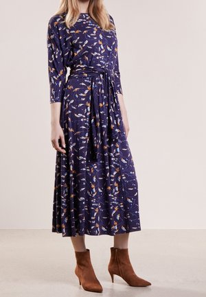 Femme portant une robe midi bleu marine à fleurs avec des manches trois-quarts et une taille nouée, associée à des bottines en daim marron.