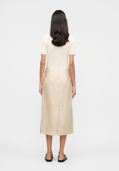 Γυναίκα με μακριά σκουρόχρωμα μαλλιά που φοράει κρεμ ριγωτό κοντομάνικο τοπ, μπεζ midi φούστα με σχίσιμο στο πίσω μέρος και μαύρα παπούτσια τύπου slip-on, στέκεται με την πλάτη προς τα εμπρός.