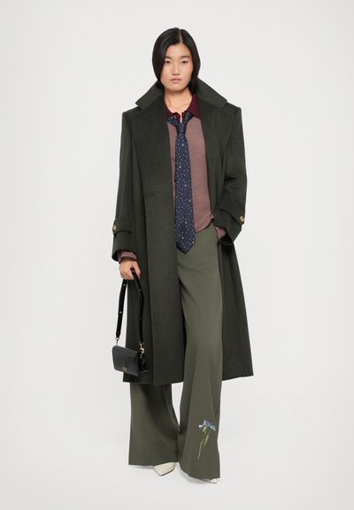 Manteau long olive, chemise bordeaux, cravate à pois navy, large pantalons olive avec broderie florale bleue, et un sac à main noir.
