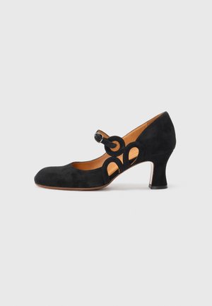 Black suede Mary Jane shoe with a rounded toe, circular buckle, and cut-out design, complemented by a mid-height heel.