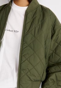 Zelenohnědá prošívaná bomber bunda s žebrovaným límcem, zipem vpředu a elastickými manžetami, vrstvená přes bílý tričko s černým textem.