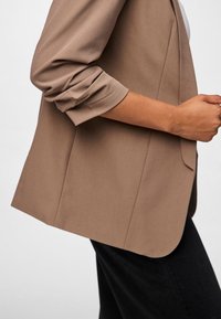 Mujer con blazer color marrón claro con las mangas remangadas y pantalones negros, vista de perfil contra un fondo liso.