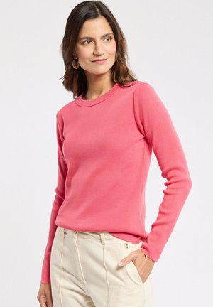 Femme portant un pull à manches longues rose et un pantalon beige clair, la main dans la poche, regardant légèrement sur le côté avec un sourire doux.