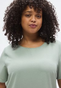 Ανοιχτό πράσινο, υφασμάτινο κοντομάνικο τοπ με στρογγυλή λαιμόκοψη, εμφανιζόμενο σε άτομο με σγουρά μαύρα μαλλιά και ουδέτερο μακιγιάζ.