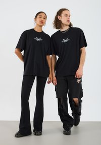 Black oversized t-shirts feature a graphic design in the center. The models wear black flared and distressed pants with black footwear.