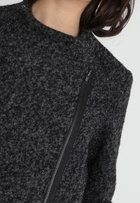 Chaqueta gris texturizada con un alto cuello, que presenta un cierre de cremallera negra en la parte frontal. La tela muestra un tejido suave y peludo.