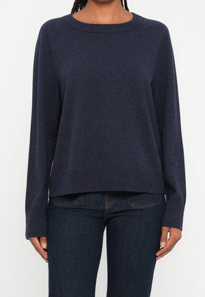 Femme portant un pull à manches longues bleu marine foncé à col rond, associé à un jean bleu foncé, debout devant un fond blanc uni.