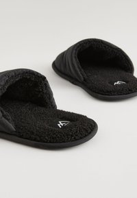 Chaussons noirs et doux avec intérieur en peluche et semelles en caoutchouc moulé. Présentent une texture extérieure matelassée et un logo sur la semelle intérieure.