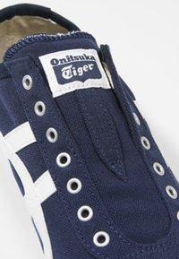 Tmavě modré plátěné nazouvačky s bílými pruhy, zpevněnými očky a výraznou nášivkou Onitsuka Tiger na jazyku. Gumová podrážka.