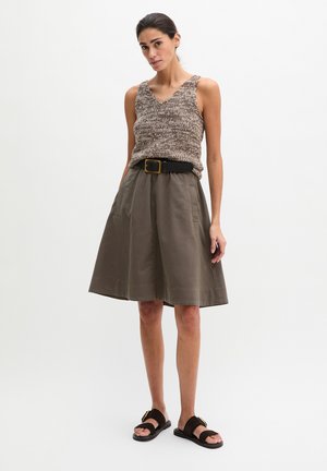 Frau steht und trägt ärmelloses Stricktop, hoch taillierten A-Linien-Rock knielang mit Gürtel und schwarze Sandalen vor weißem Hintergrund.