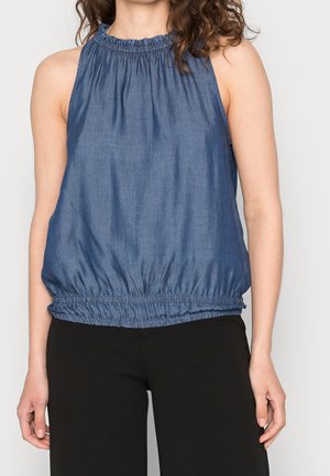 Femme portant un haut bleu sans manches avec un col froncé et un ourlet élastique, associé à un pantalon noir, debout devant un fond uni.