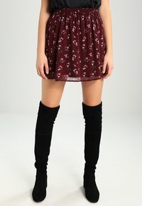 Falda de flores burdeos con cintura elástica, en capas, de textura translúcida, que muestra pequeños patrones florales coloridos. Combinada con botas negras por encima de la rodilla.