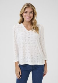 Haut à manches longues blanc avec un design à œillets, décolleté en V et coupe décontractée. Fabriqué en tissu léger, il a une texture douce et des poignets roulés.