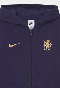 Sudadera con cremallera color marino hecha de tela suave, con un logo dorado de Nike en el pecho izquierdo y un escudo dorado de un león en el derecho.