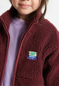 Burgundska flis jakna s teksturiranim završetkom, zatvaranje na zip, visoki ovratnik i šareni patch s natpisom "LA MAISON AMV SUPER VINTAGE."