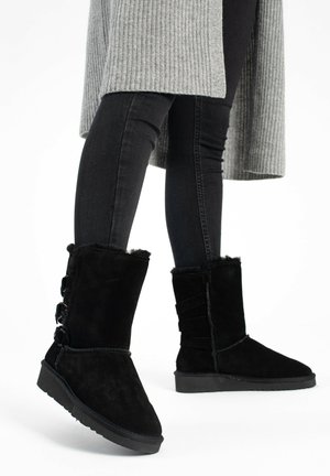 Black suede ankle boots with a flat sole, featuring plush lining, button accents on the side, and gathered texture at the back.
