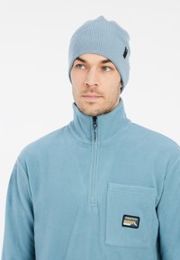 Šviesiai mėlynas fliso pusiau užtraukiamas palaidinukas turi priekinį krūtinės kišenę su logotipo aplikacija. Atitinkantis megztas skrybėlė turi raštuotą tekstūrą.