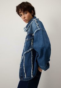 Jauns cilvēks ar īsiem cirtainiem matiem, uzvilcis pārāk lielu zilu džinsa jaku ar nodilušām vīlēm, stāv uz vienkārša gaiša fona.