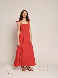Mulher com cabelo encaracolado e comprido, vestindo um vestido maxi sem mangas vermelho, com alças amarradas e bainha plissada, em pé num chão coberto de confetes.