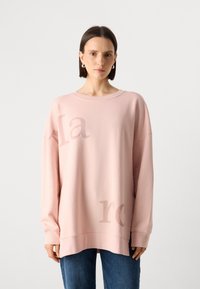 Svetloružová oversized mikina z jemného materiálu s širokým výstrihom a spustenými ramenami. Na prednej strane je diskrétny potlač textu.