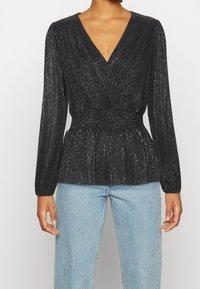 Blusa avvolgente nera a maniche lunghe con scollatura a V profonda, tessuto texturizzato e scintille argentate; vita cinte; abbinata a jeans in denim azzurro chiaro.
