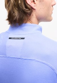 Άτομο με ανοιχτά καστανά μαλλιά που φοράει ένα υφασμάτινο ανοιχτό μπλε πουκάμισο με μια μαύρη ετικέτα στην πλάτη που γράφει «All Conditions Gear».