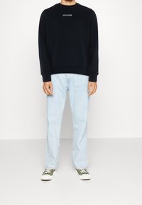 Felpa blu marino con logo ricamato bianco, abbinata a jeans in denim azzurro chiaro e scarpe da ginnastica in tela verde con suola bianca.