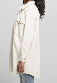 Femme portant une surchemise en velours côtelé blanc oversize avec poches poitrine boutonnées et poignets, associée à un jean bleu clair, vue de profil.
