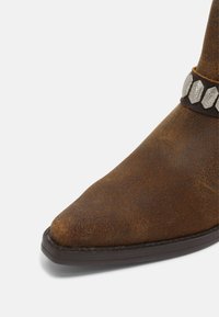 Botte de cowboy en daim marron avec un bout pointu et une semelle foncée, équipée d’une lanière en cuir ornée de clous hexagonaux argentés autour de la cheville.