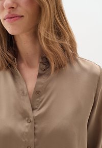 Blusa in satin marrone con chiusura anteriore a bottoni, colletto e texture liscia. Presenta piccoli bottoni marroni lungo la piastrella.