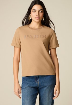 Žena s dlouhými tmavými vlasy má zlaté abstraktní náušnice, opálené tričko s nápisem "PARIS" a modré džíny, stojící před neutrálním pozadím.