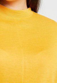 Top amarillo de cuello alto con canalé, hecho de una tela suave; presenta un diseño ajustado con costuras mínimas y una textura suave.