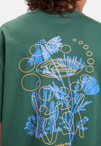Camiseta de algodón verde con un diseño gráfico en azul y naranja, que representa figuras abstractas y elementos de plantas en la parte posterior.