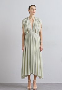 Rochie lungă, fluidă, din material verde deschis, cu plisuri, decolteu adânc și umeri drapati. Asortată cu pantofi cu toc nude.