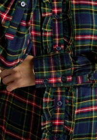 Plaid shirt in dark blue, green, red, and yellow hues. Features ruffles along the button placket and navy buttons. Cuffed sleeves visible.
