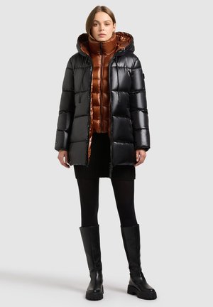 Abrigo negro acolchado con capucha, superpuesto a una chaqueta acolchada marrón. Ambos tienen un acabado brillante; la modelo lleva mallas negras y botas hasta la rodilla.