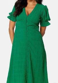 Vestido verde de botones con mangas cortas abullonadas, que presenta una tela texturizada y un escote en V. La falda fluye hasta la altura de la mitad de la pantorrilla.