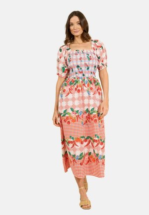 Žena v midi šatách s nafúknutými rukávmi, naškrobeným korzetom a vzorom červeného ovocia a kvetov, stojaca pred jednoduchým pozadím.