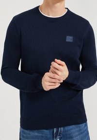 Man med en marinblå stickad tröja med en liten "Polo Club"-logotyp, matchad med blå jeans, med händerna knäppta framför sig.