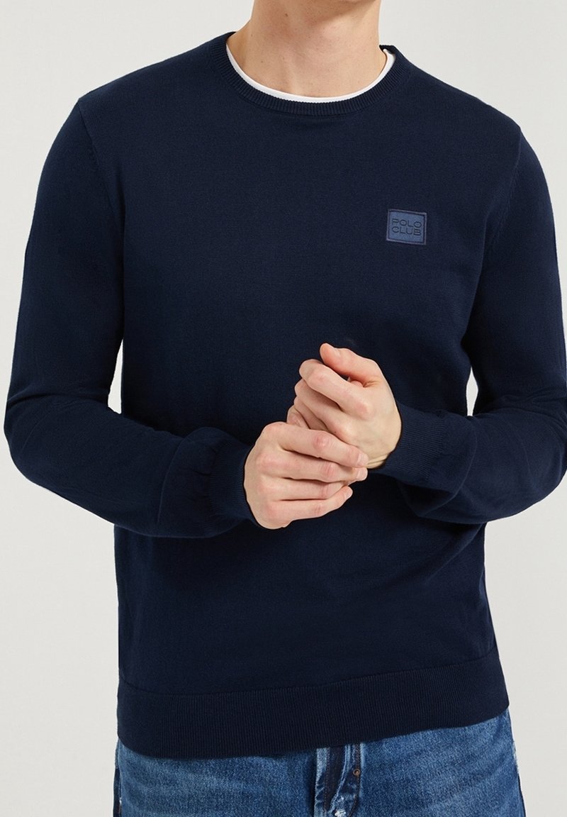 Man med en marinblå stickad tröja med en liten "Polo Club"-logotyp, matchad med blå jeans, med händerna knäppta framför sig.