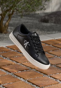 Svart lädersneaker med vit gummisula, med snörning och Calvin Klein-logga. Positionerad ovanför en yta av trä och grus.